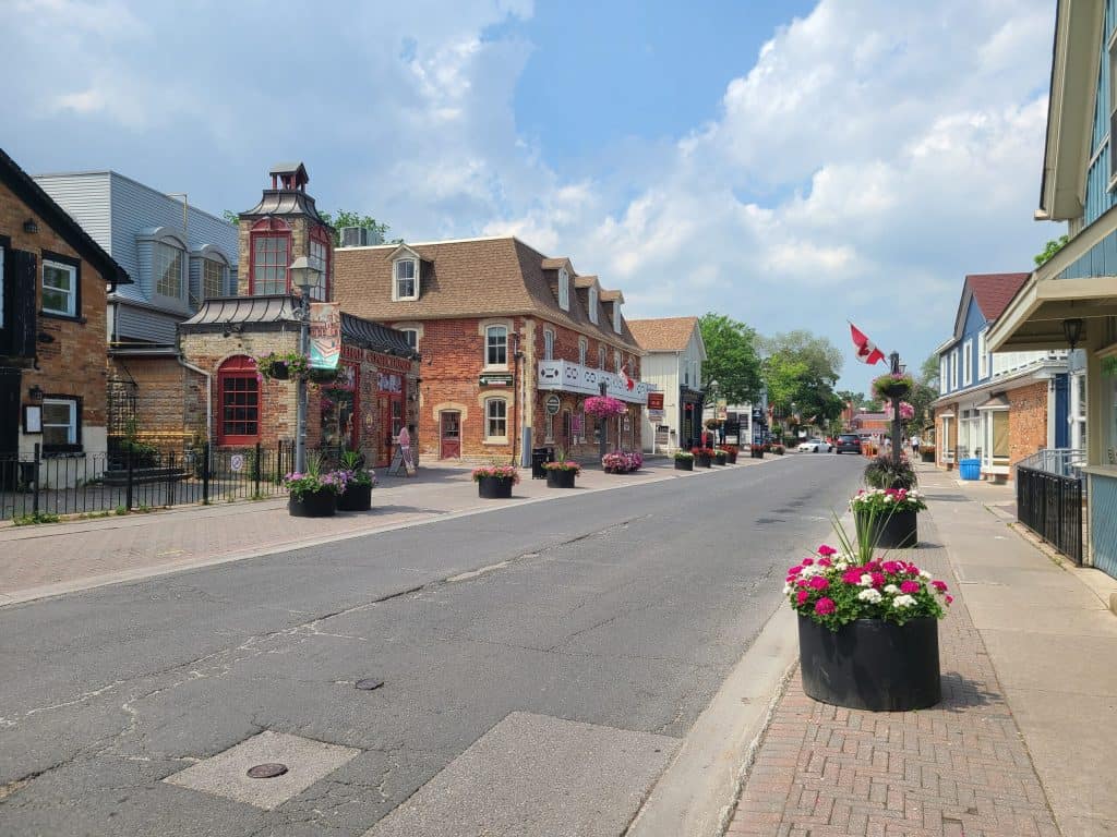 Colorful small-town street with charming historic buildings, vibrant flower arrangements in large planters, and Canadian flags, showcasing a welcoming, picturesque neighborhood scene.